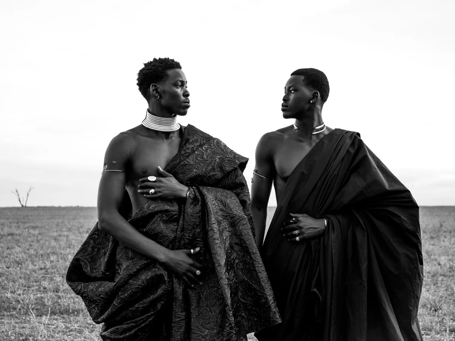 Two men draped in dark fabric face each other in an open field, in black and white.