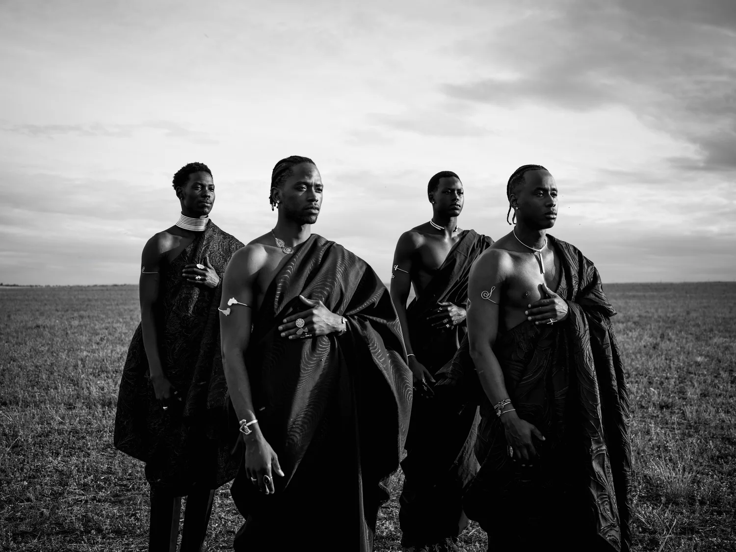 Four men wrapped in dark draped cloth stand together in a grass field.