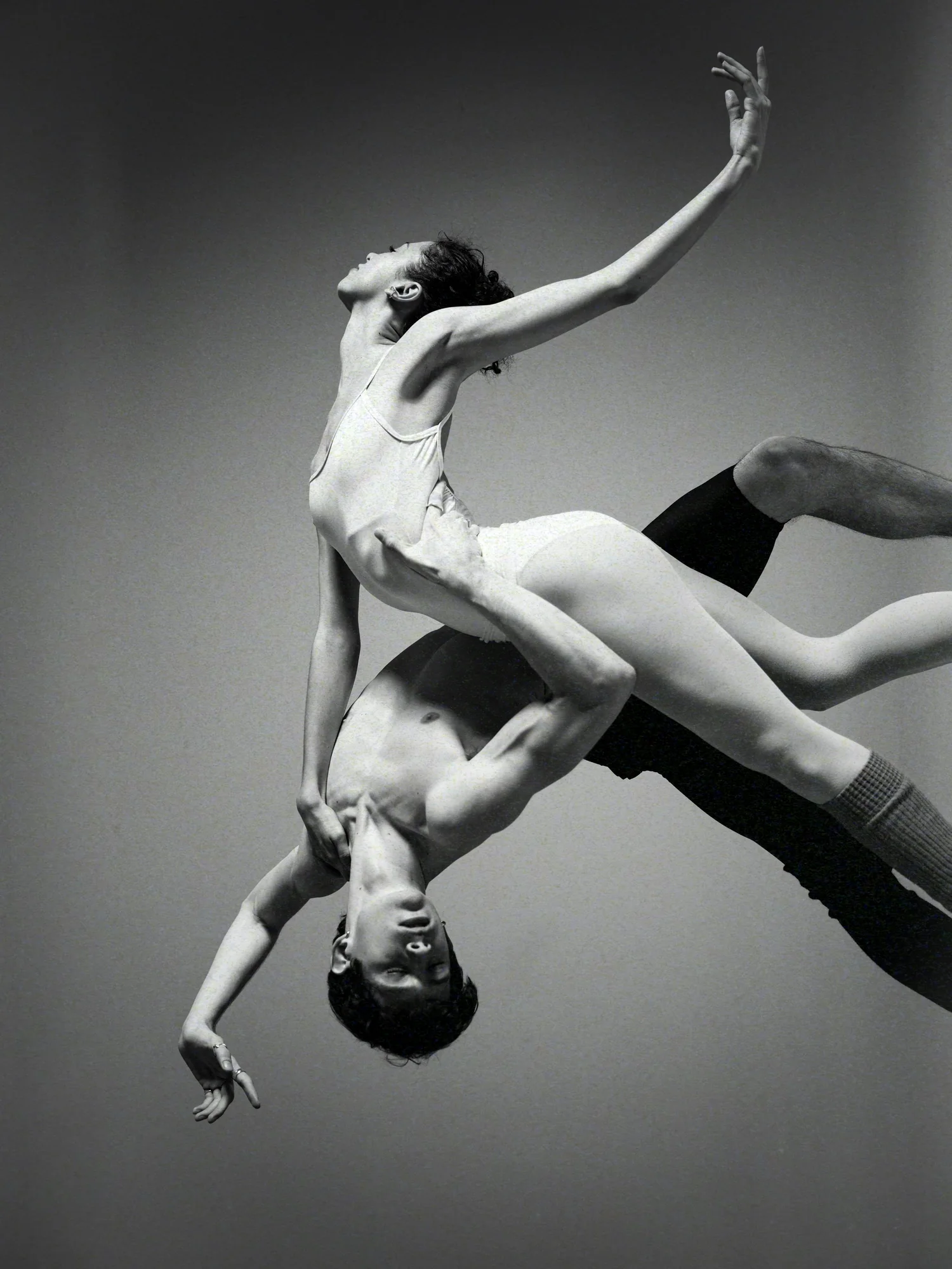 Two dancers intertwined in mid-air against a grey backdrop, one reaching upward.