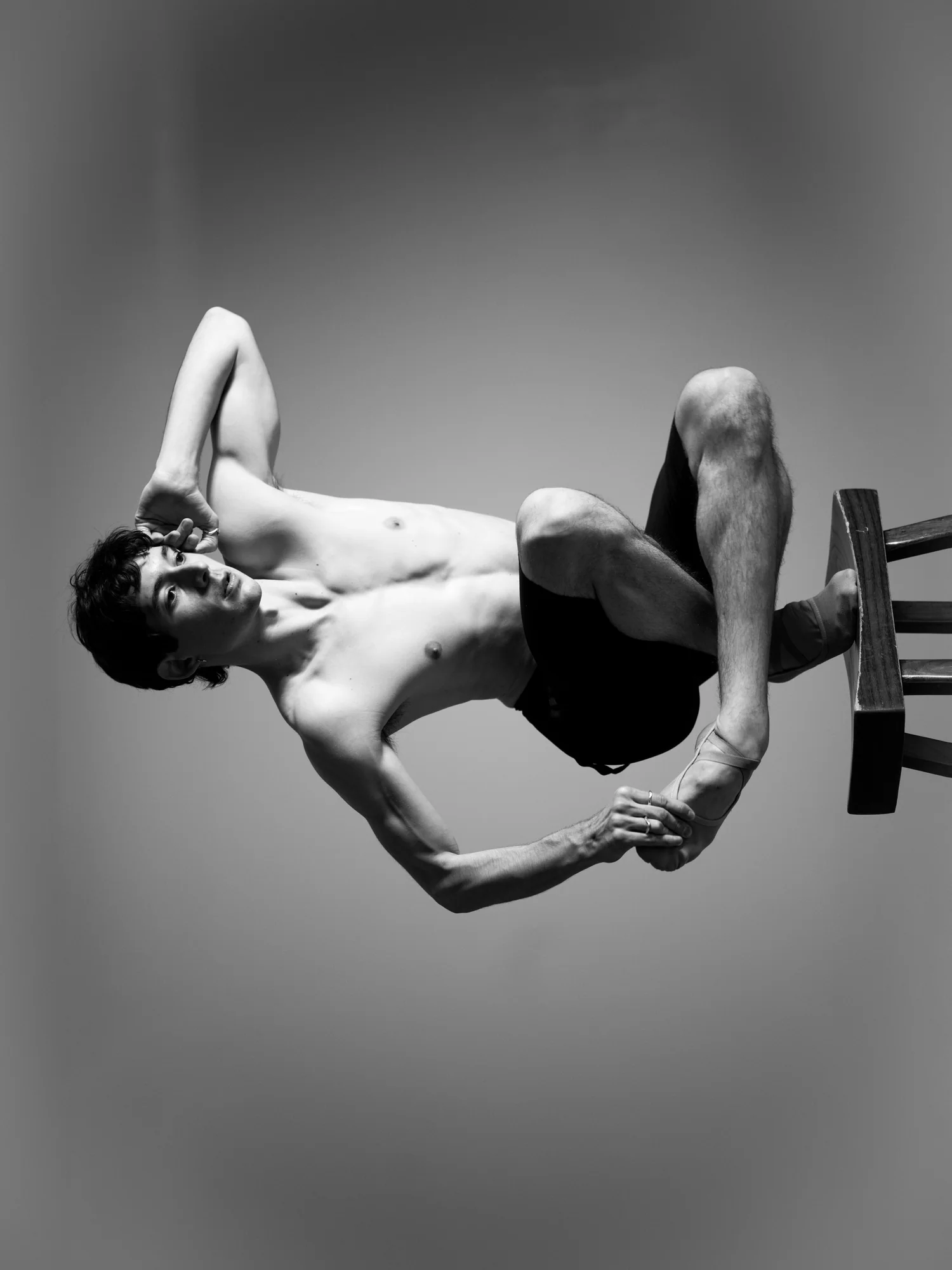 A sideways frame of a shirtless dancer crouched on a wooden stool.