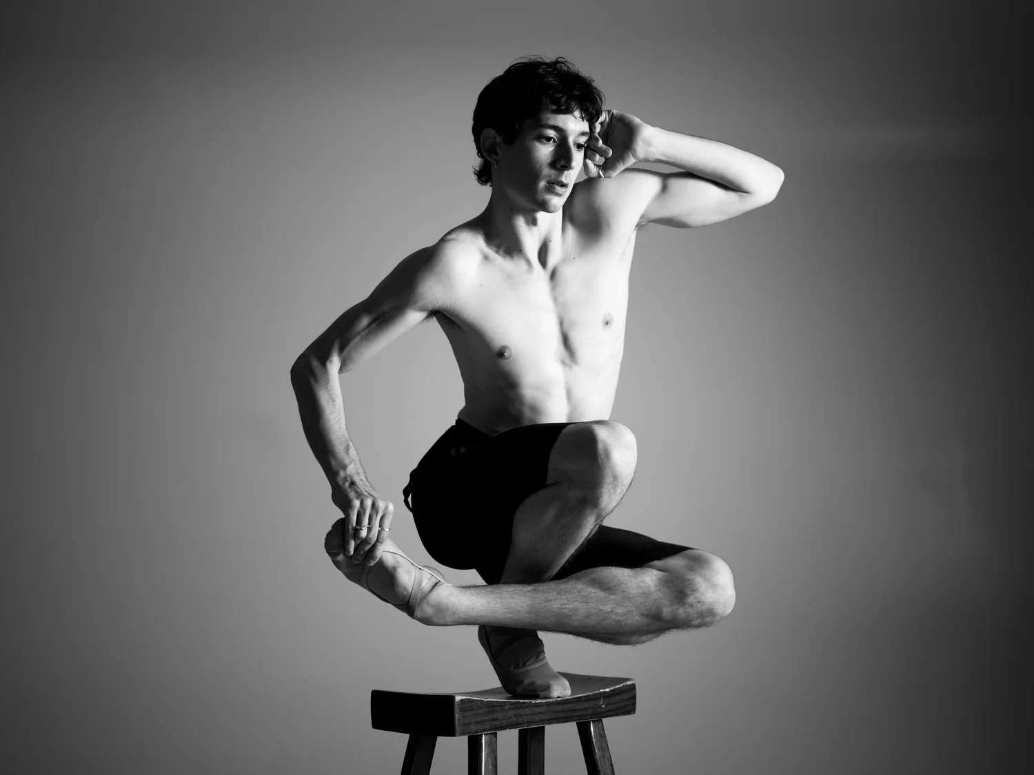 A shirtless dancer balances in a crouch on a wooden stool.