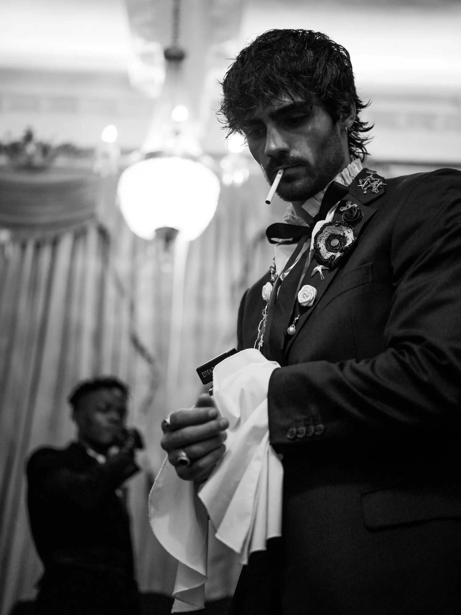 A man with a cigarette in his mouth wipes his hands on a white cloth beneath a chandelier.
