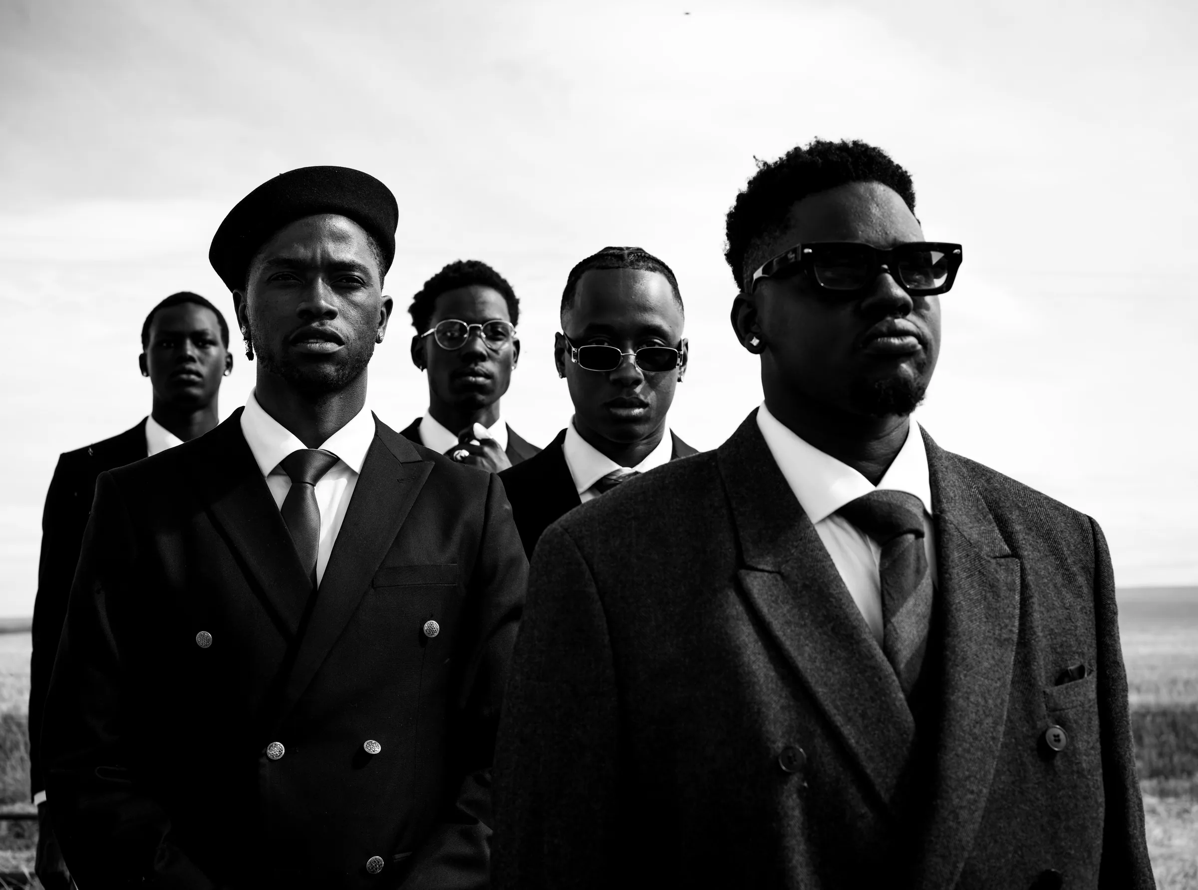 Five men in dark suits and ties stand close together under an open sky, in black and white.
