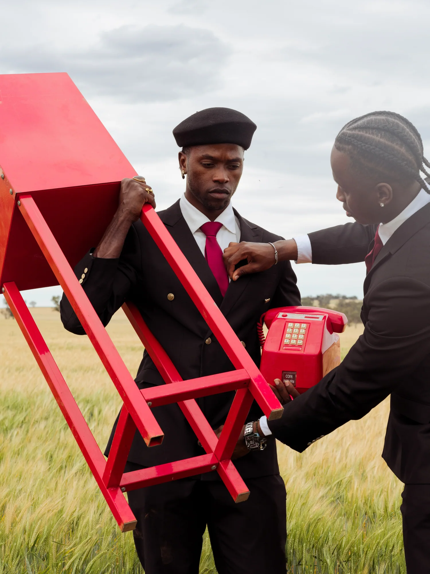 A man in a beret and suit holds a red table and a red telephone while another man straightens his tie.