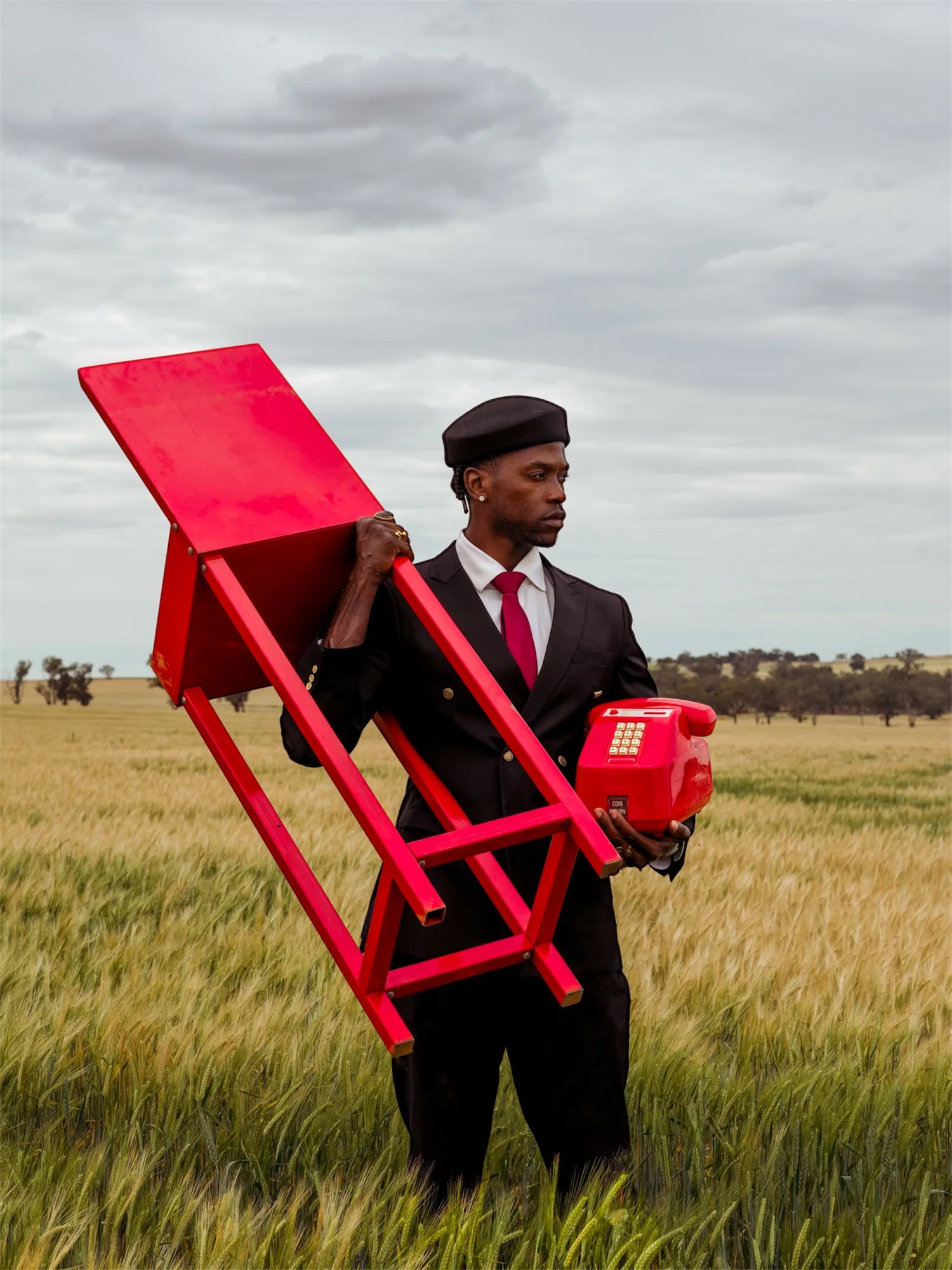 A man in a beret and dark suit carries a red table and a red telephone through a wheat field.
