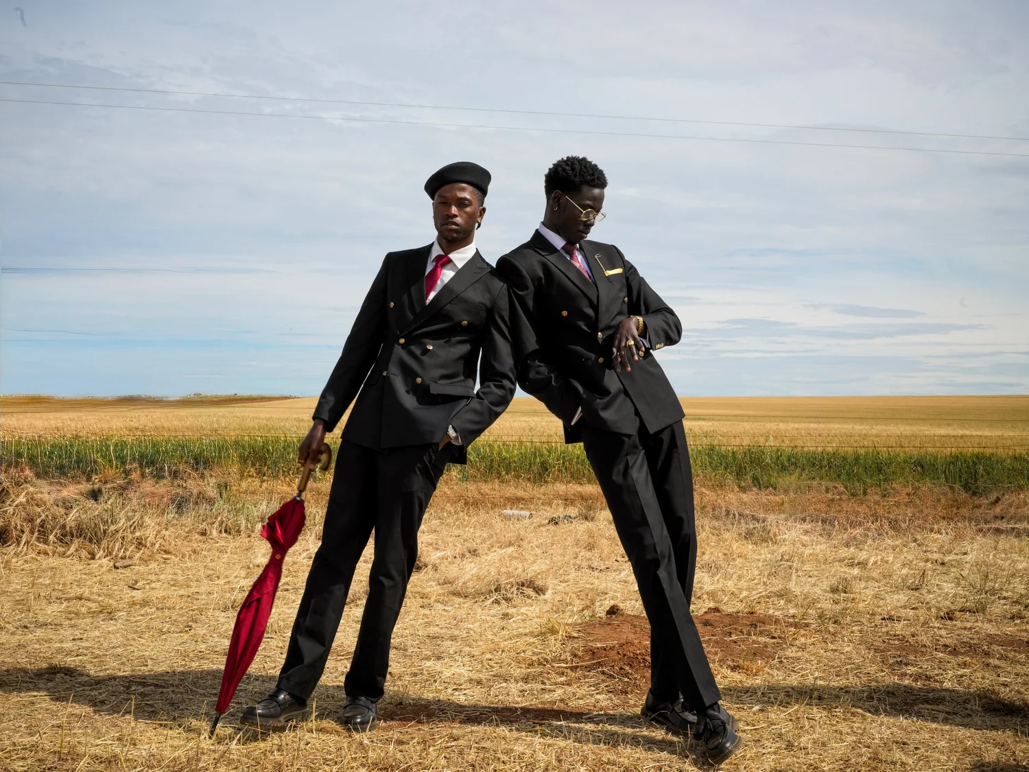 Two men in dark suits lean against each other in a dry field, one resting on a red umbrella.