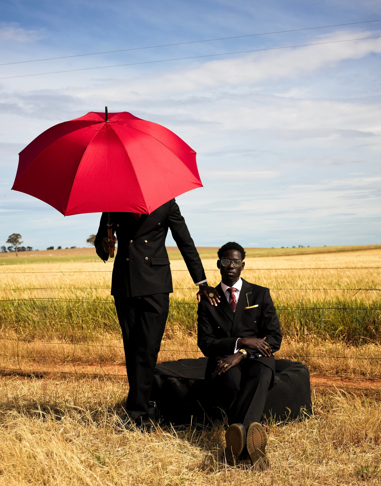 A man in a suit sits on a suitcase in a golden field while another stands behind him, his face hidden by a red umbrella.