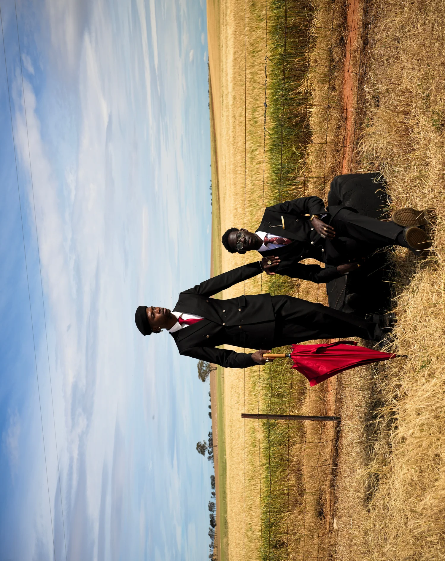 A sideways frame of two men in suits in a dry field, one standing with a red umbrella and one seated.