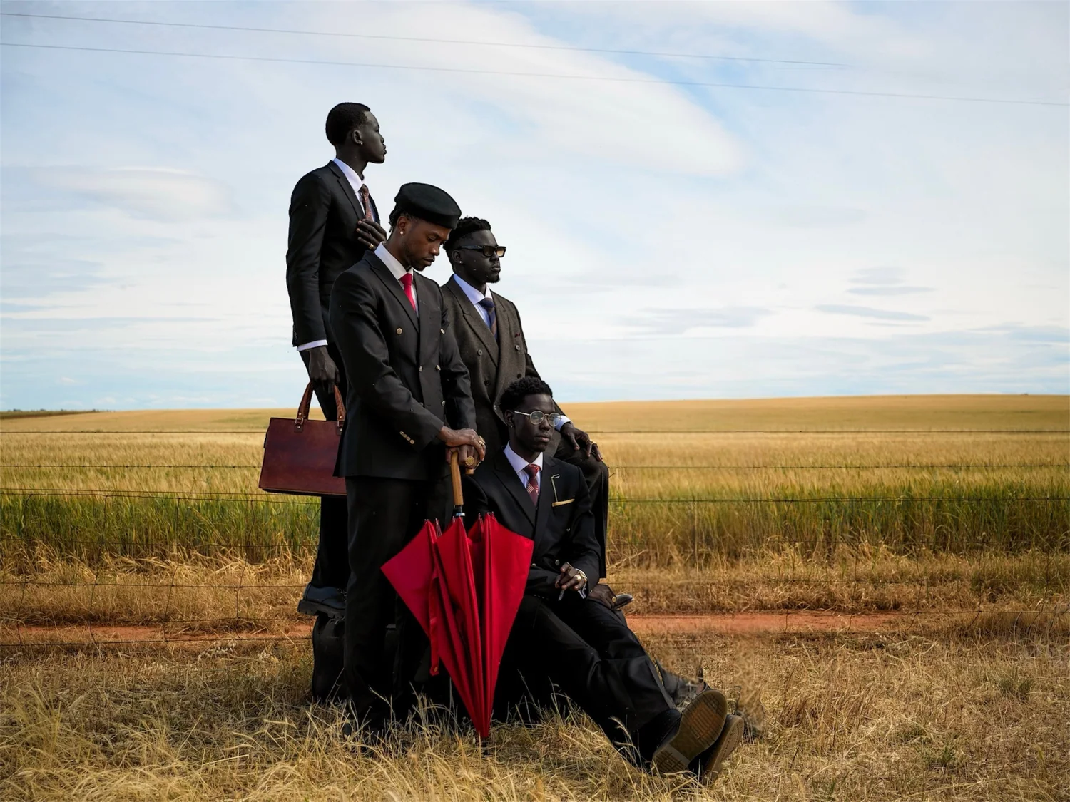 Four men in dark suits grouped in a wheat field with a red umbrella and a brown briefcase.
