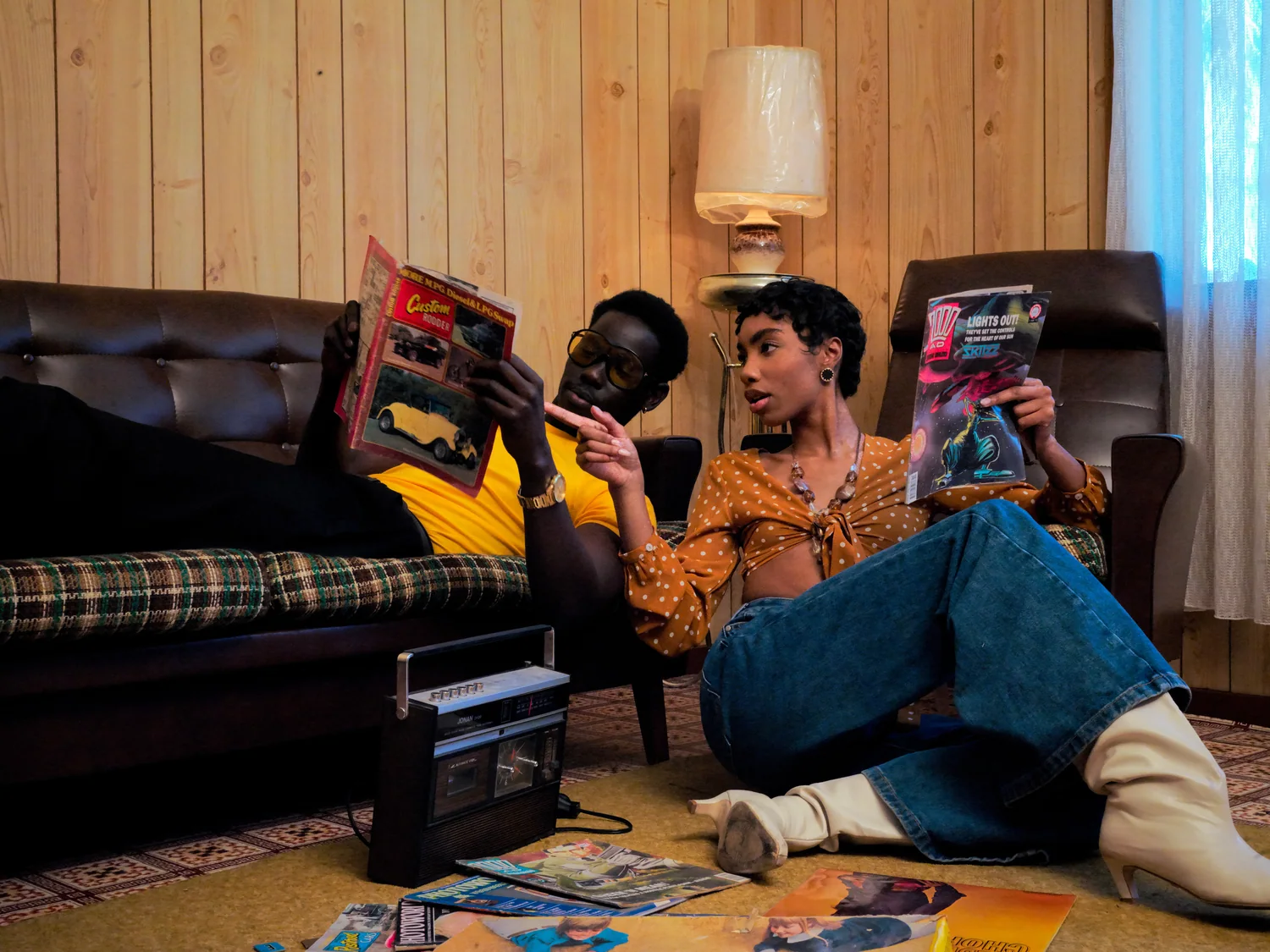 A man and a woman read magazines on a patterned carpet beside a cassette radio.