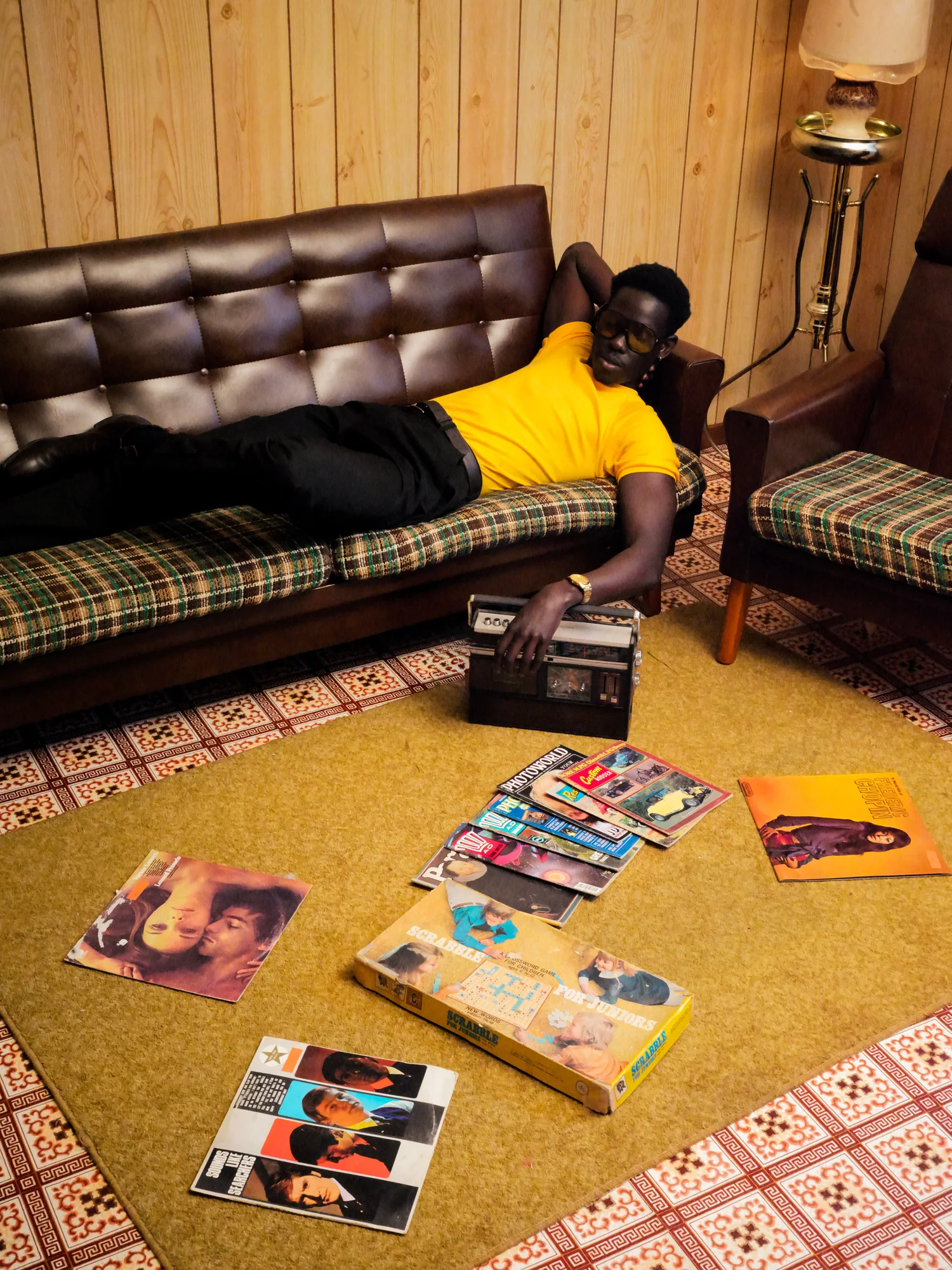 A man in a yellow shirt lies on a leather sofa reaching for a radio, magazines scattered across the carpet.