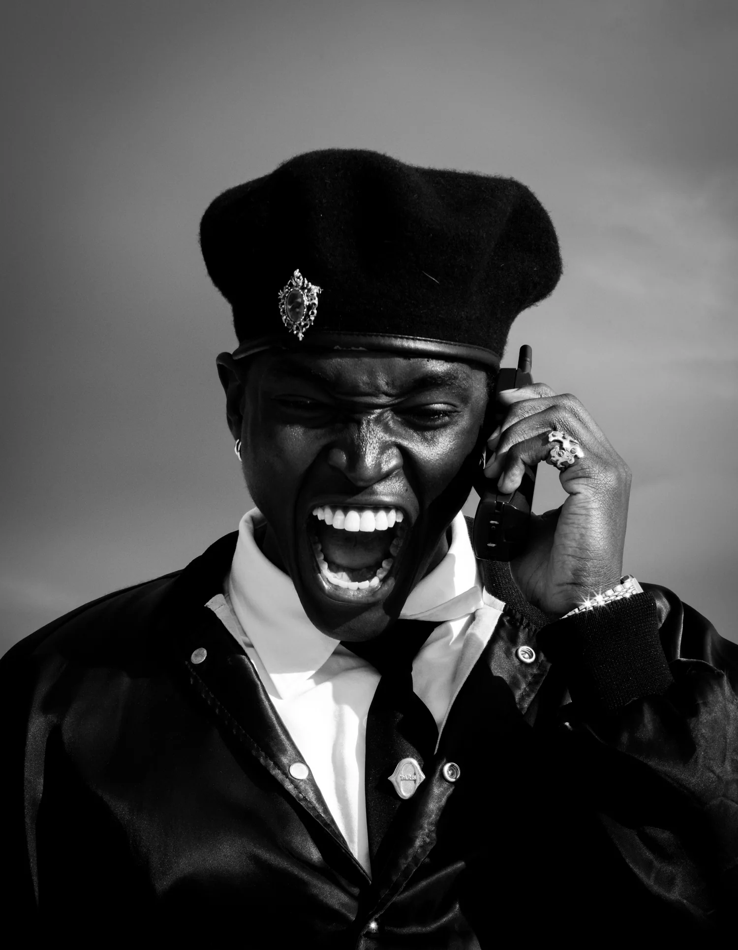 A man in a beret and leather jacket shouts into a mobile phone, in black and white.
