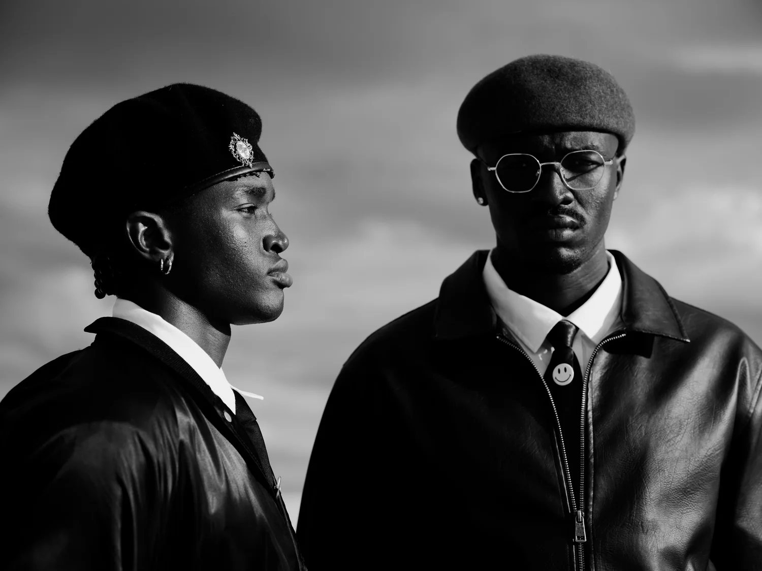 Close portrait of two men in berets and leather jackets, one in profile and one in round glasses.
