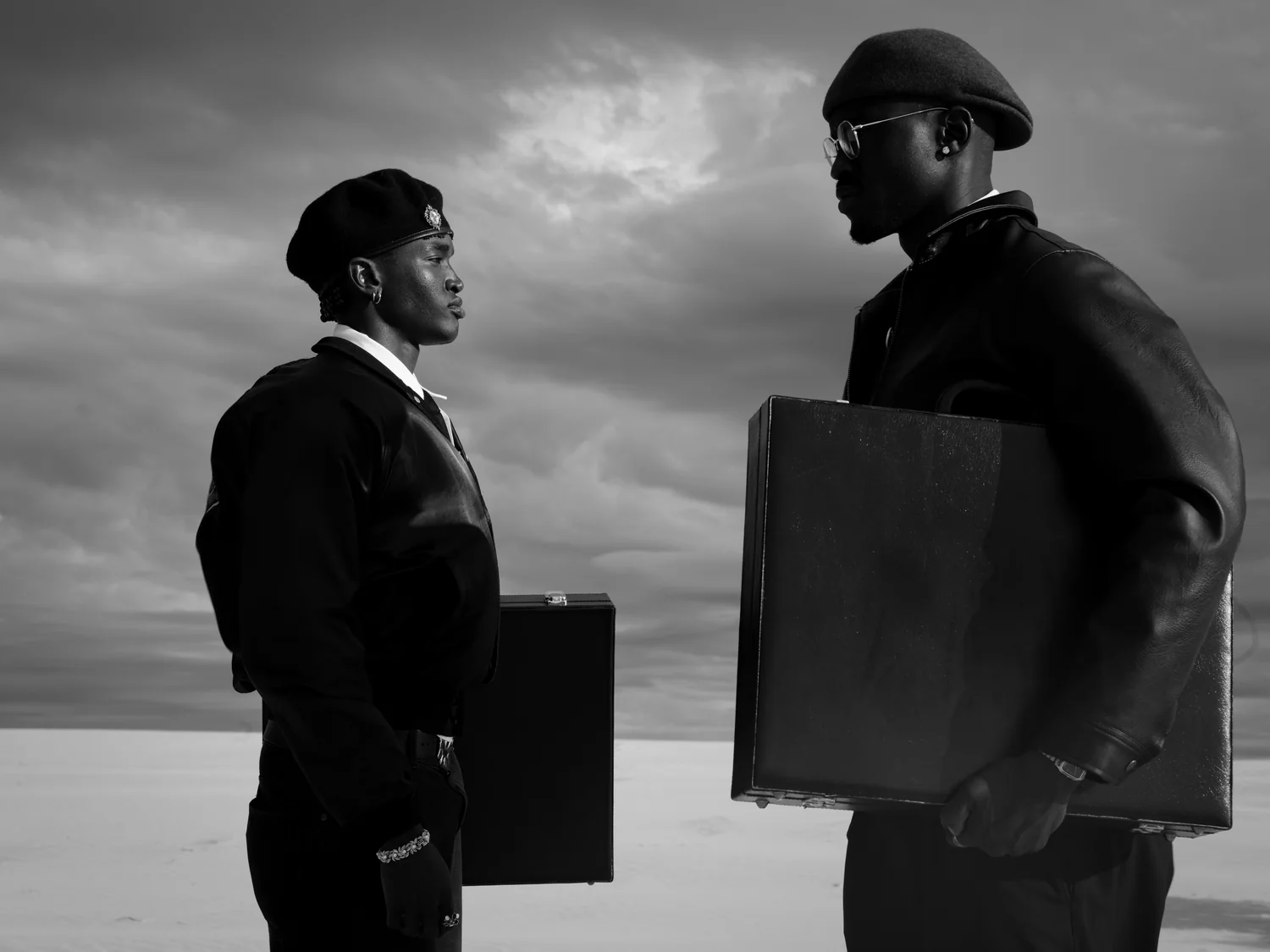 Two men in berets face each other holding briefcases under a heavy sky.