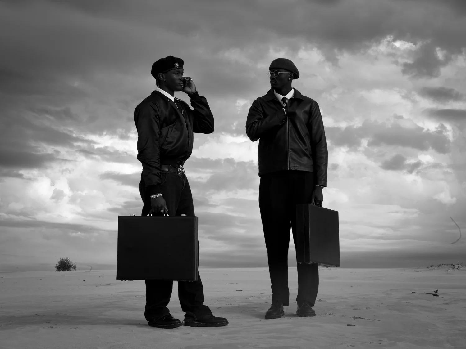 Two men in leather jackets stand on a pale, flat plain with briefcases, one of them on the phone.