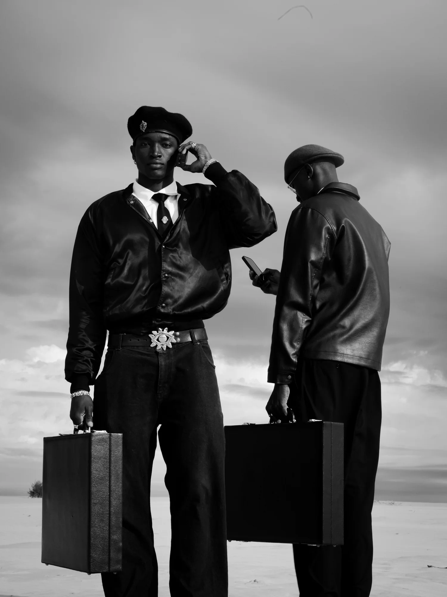 Two men with briefcases stand on a salt flat, one on the phone and one looking at his.