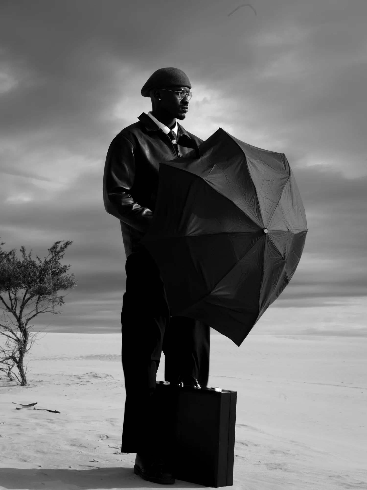 A man in a beret shelters behind a black umbrella on a salt flat, a briefcase at his feet.