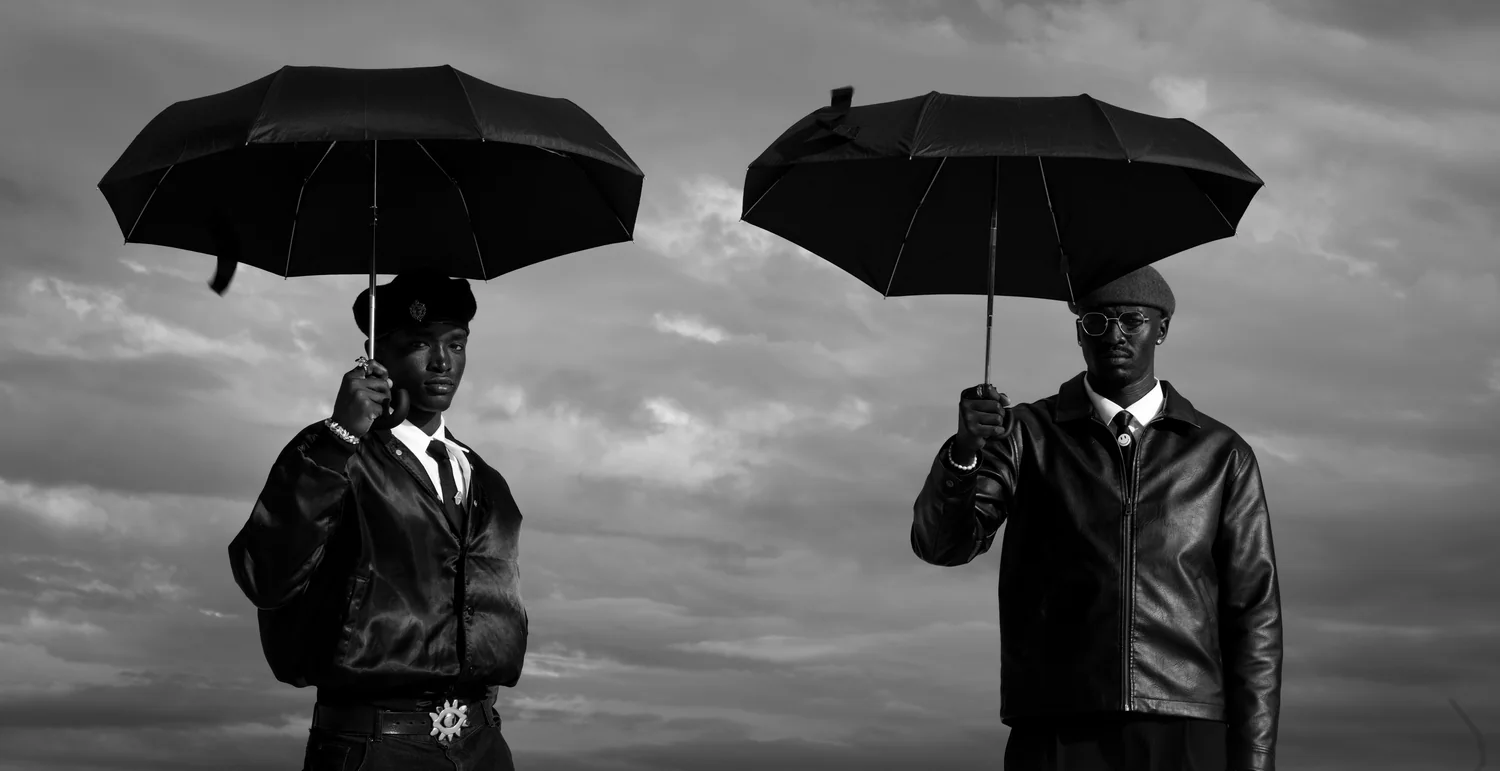 Two men in leather jackets hold black umbrellas against a clouded sky.