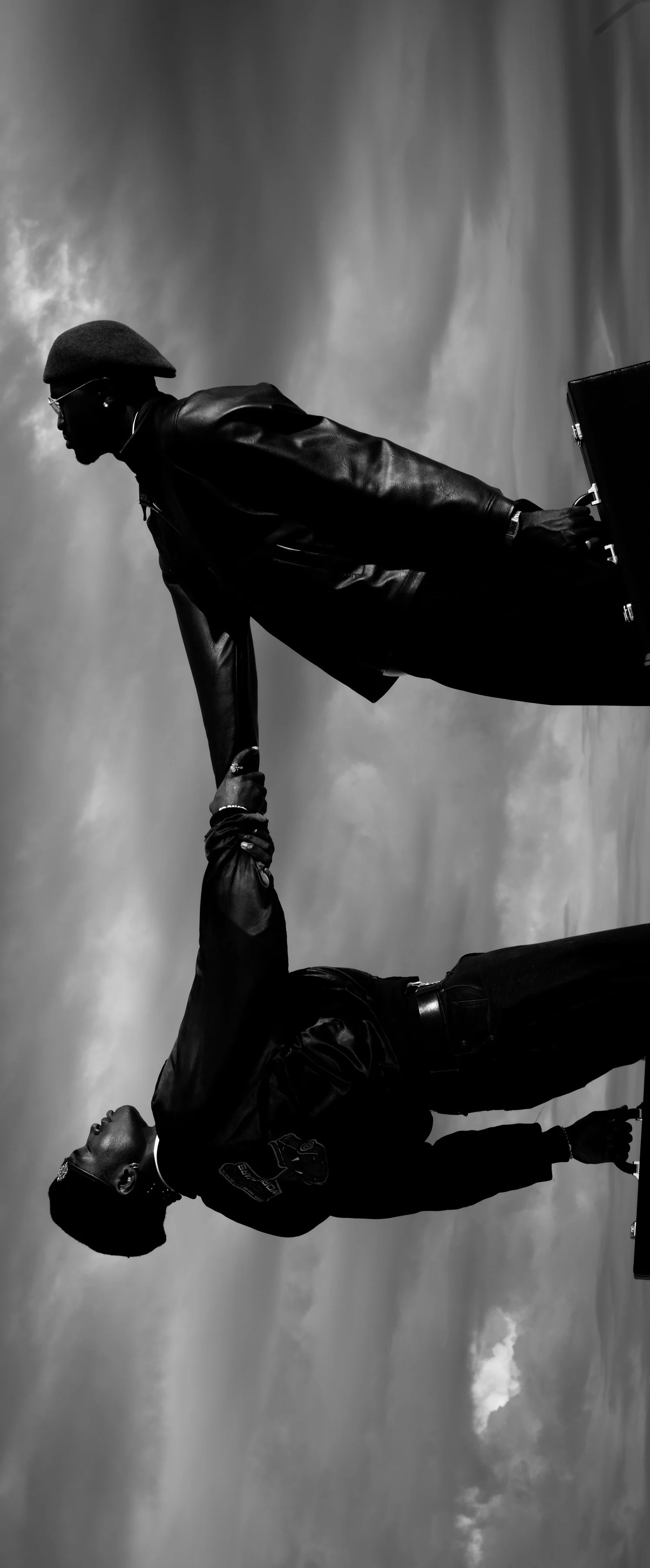 A sideways frame of two men in leather jackets leaning away from each other, hands clasped, against the clouds.