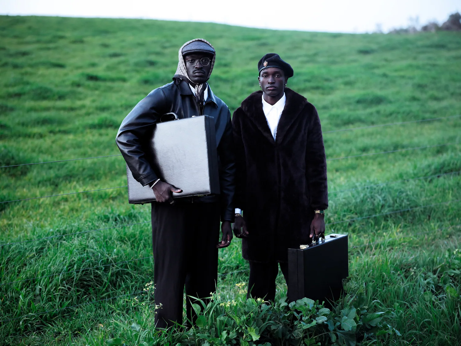 Two men, one in a leather jacket and one in a fur coat, stand in a green field holding briefcases.