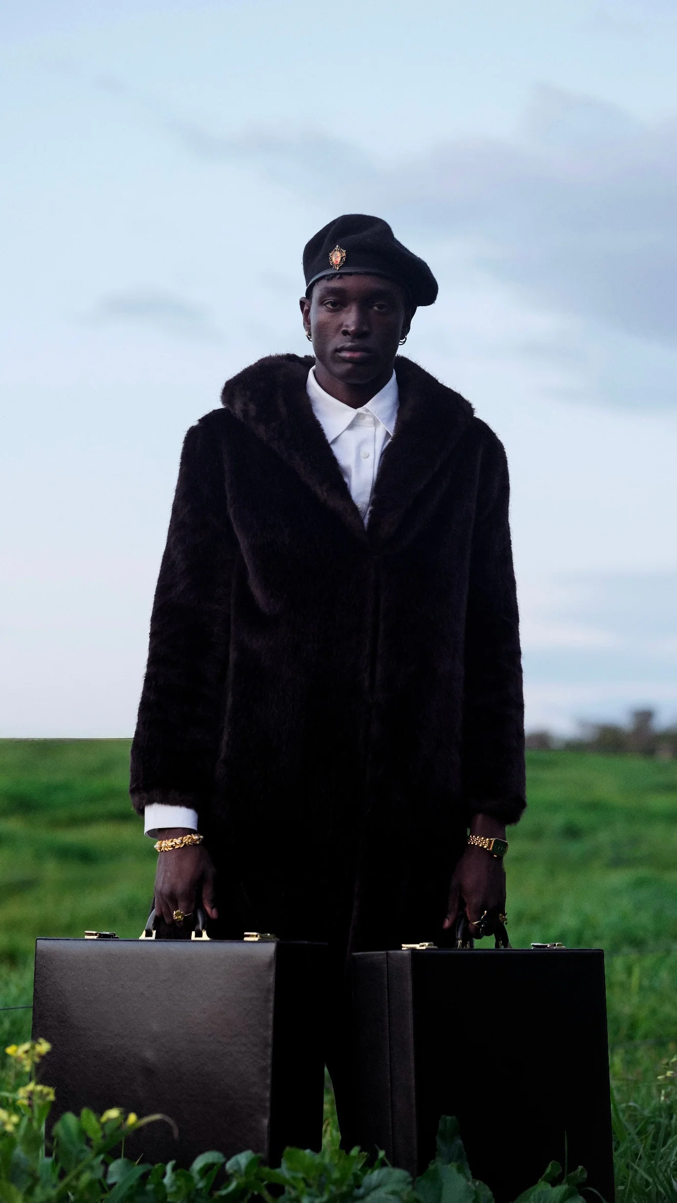 A man in a beret and a black fur coat stands in a green field with a briefcase in each hand.