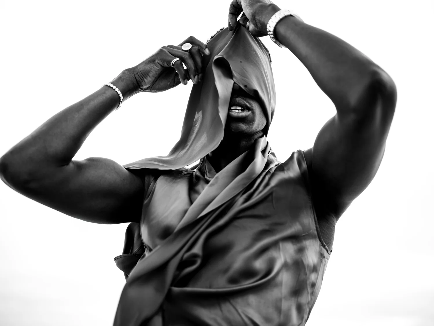 A man lifts satin fabric over his face with both arms raised, in black and white.