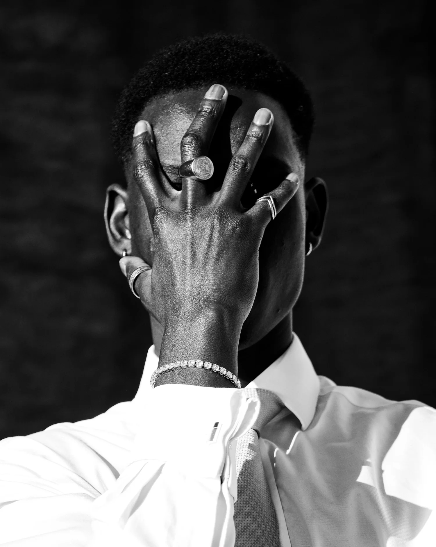 A man hides his face behind a hand wearing silver rings, in black and white.