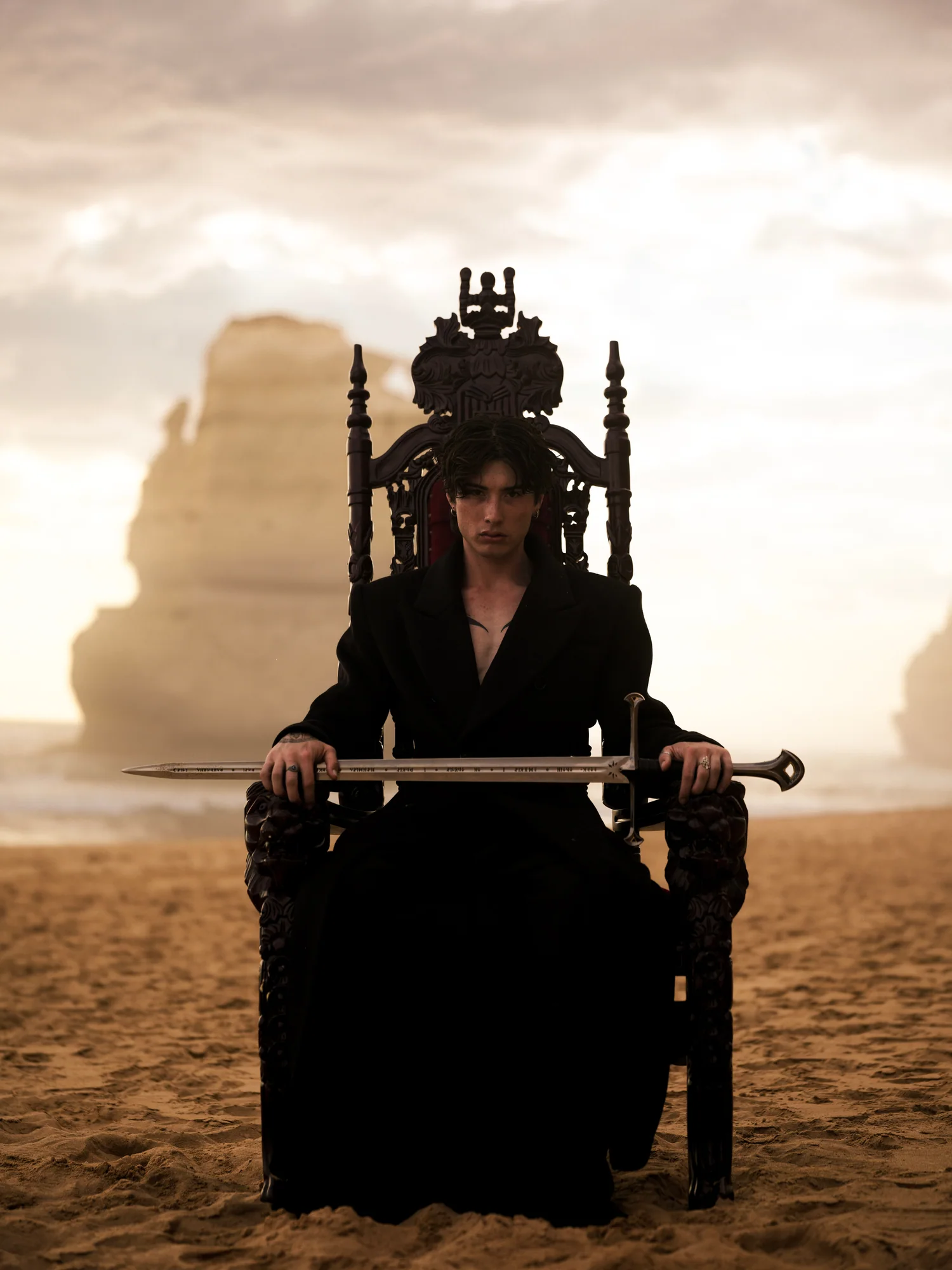 A figure in a black coat sits on a carved throne on a beach with a sword across their lap.