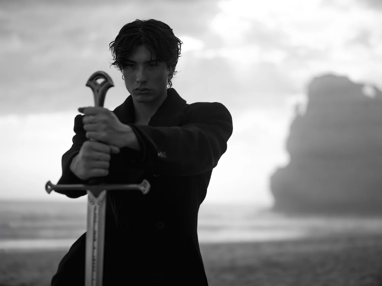 A man in a dark coat rests both hands on a sword planted in front of him, a sea stack behind, in black and white.