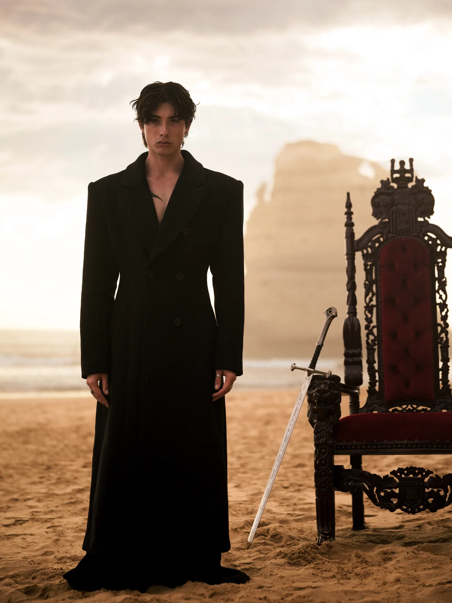 A man in a long black coat stands on the sand beside a carved throne and a sword.