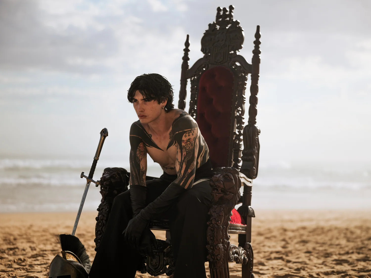 A man with tattooed arms sits forward on a carved throne on the beach.
