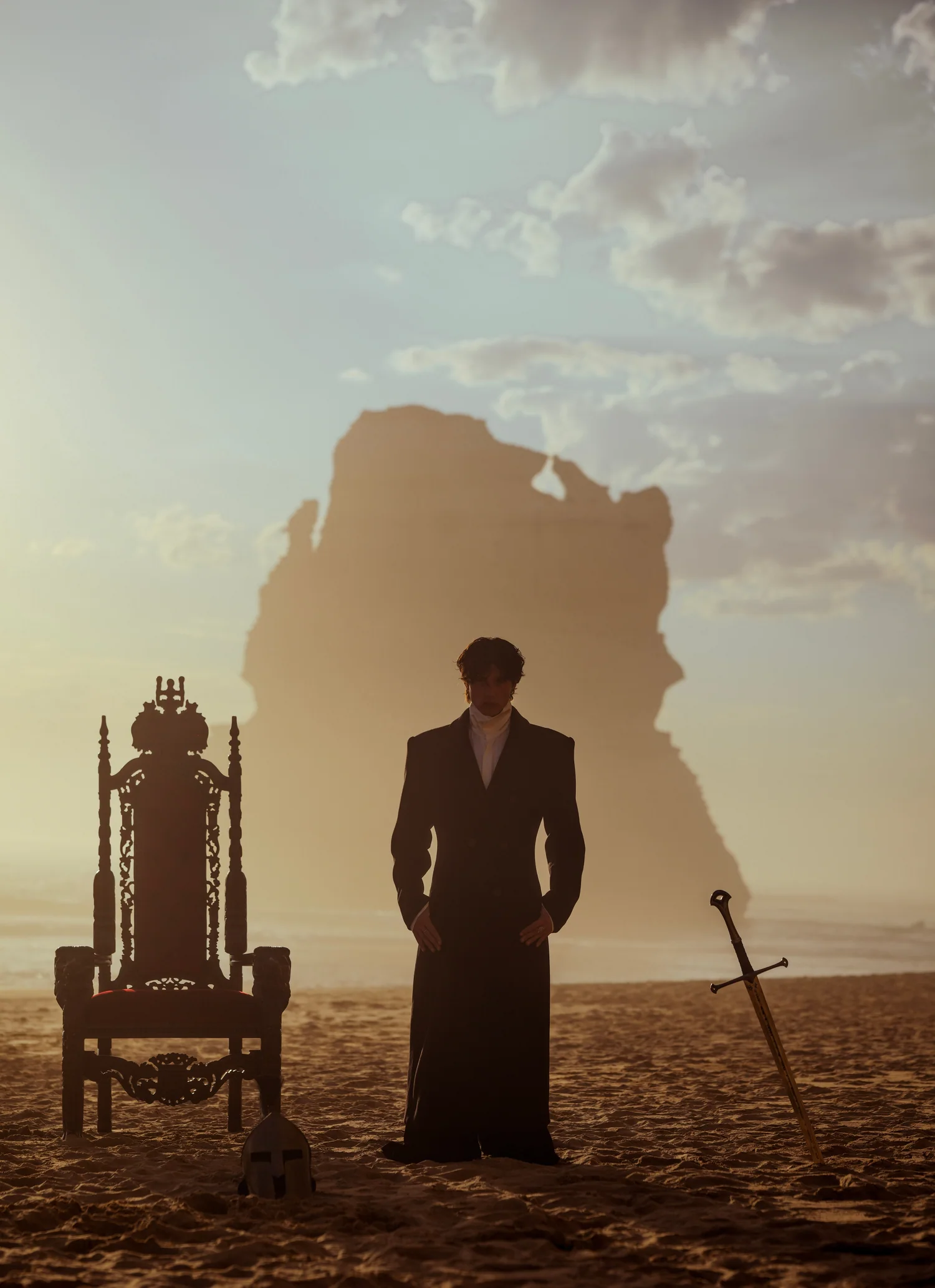 A lone figure in a long coat stands between a throne and a sword in the sand at dusk.